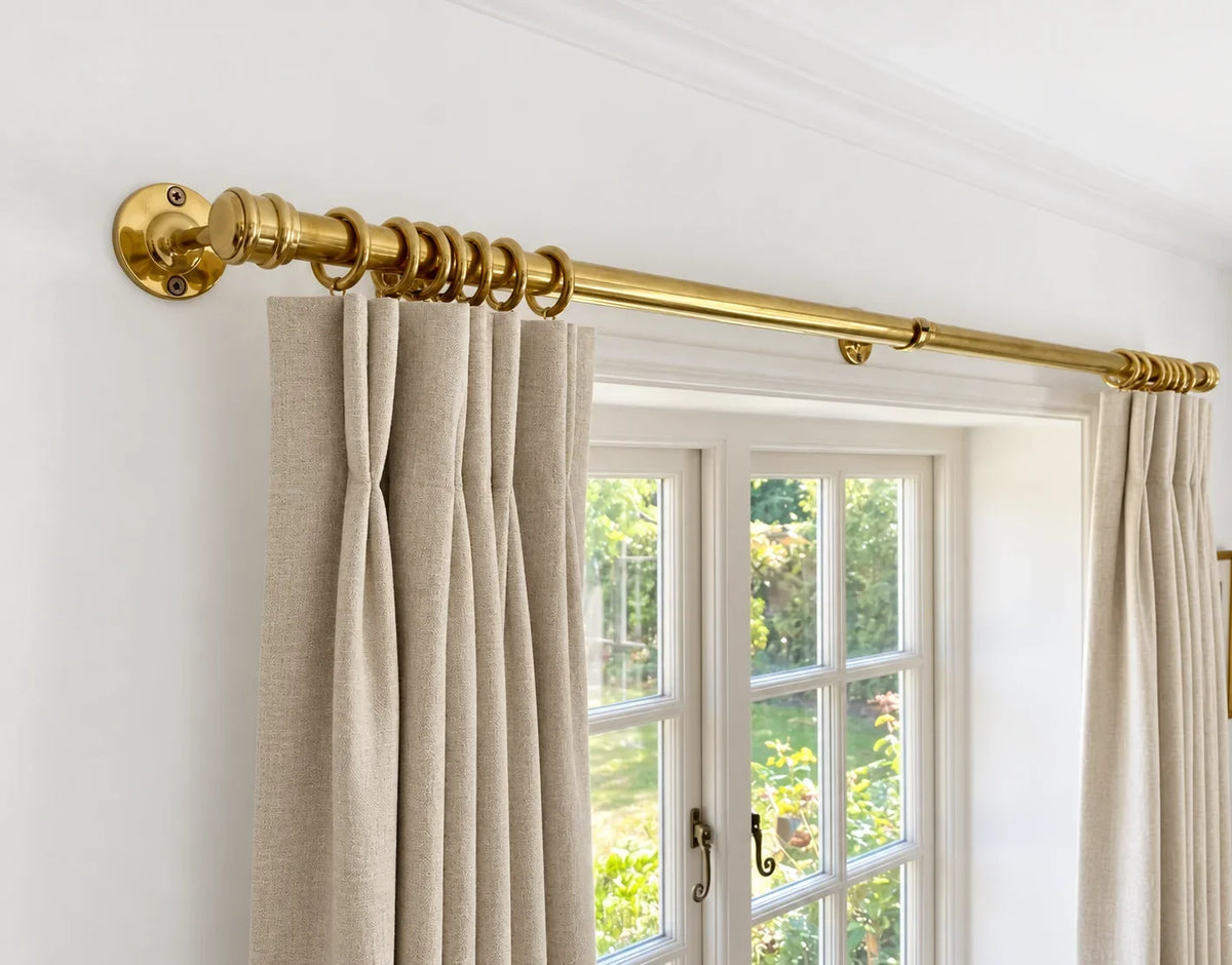 A brass curtain rod with matching rings mounted on a white wall above a window, styled with beige linen curtains.