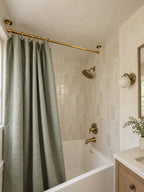 A modern bathroom featuring a sage green linen shower curtain, a brass shower rod, matching brass showerhead and faucet, and a white tiled wall.