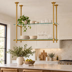 A modern brass double-shelf hanging kitchen island rack with clear glass shelves, styled with dishes and plants in a bright, contemporary kitchen.