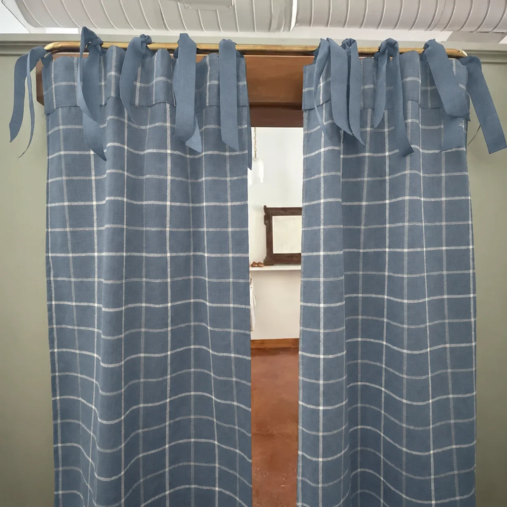 A pair of dusty blue window curtains with a white windowpane plaid pattern, hanging on a wooden rod with tie-back tabs.