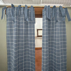 A pair of dusty blue window curtains with a white windowpane plaid pattern, hanging on a wooden rod with tie-back tabs.