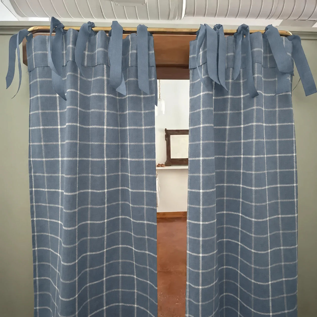 A pair of dusty blue window curtains with a white windowpane plaid pattern, hanging on a wooden rod with tie-back tabs.