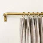 A close-up of light beige pleated curtains hanging from a brass curtain rod with matching rings against a plain off-white wall.