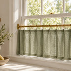 A sage green linen window valance hanging from a brass rod on a white window frame, with a bright outdoor view in the background and a decorative bowl of pears on the windowsill.