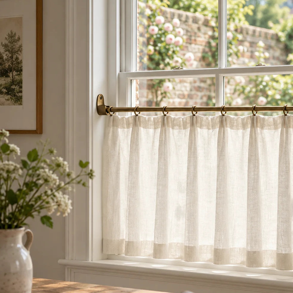 A sheer off-white curtain panel hanging from a brass rod on a white window in a bright kitchen or dining room, with a wooden table, chair, and vase of flowers visible in the foreground.